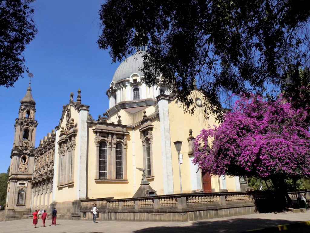 This photo shows Holy Trinity Cathedral set against a brilliant blue sky with vibrant purple blossom in the foregraound