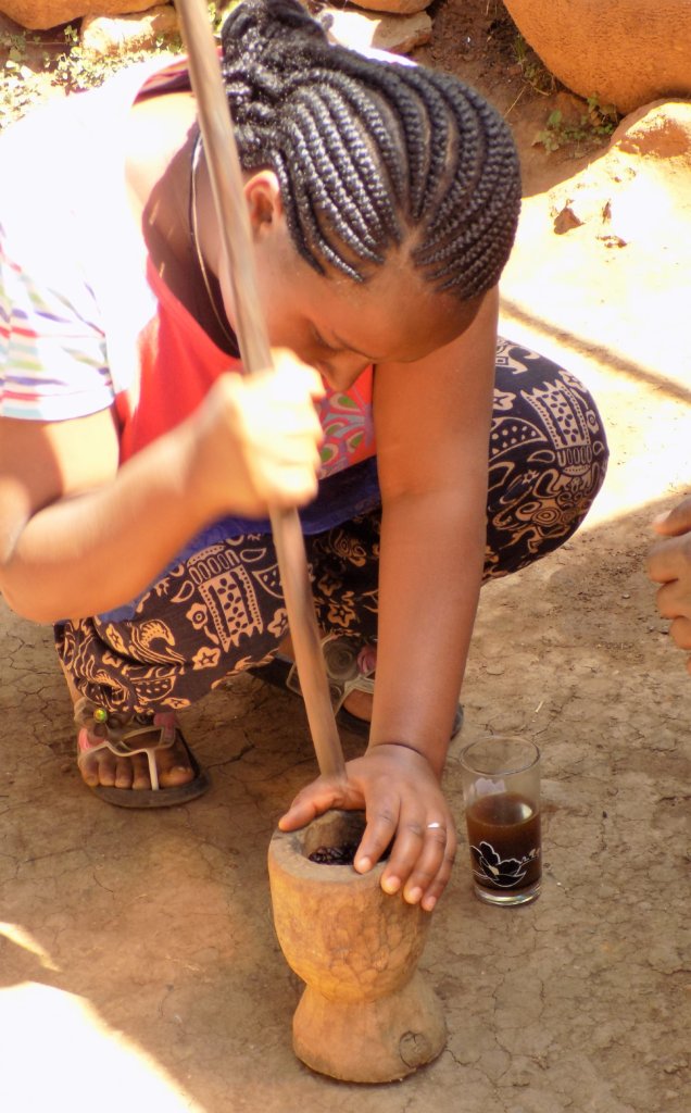 This photo shows a lady crouching on the ground grinding coffee beans in a pestle and mortar