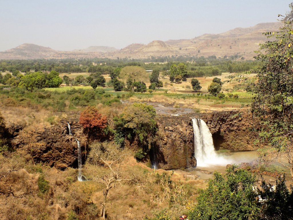 This photo shows a wide view of the Blue Nile Falls set against a backdrop of verdant countryside and mountains