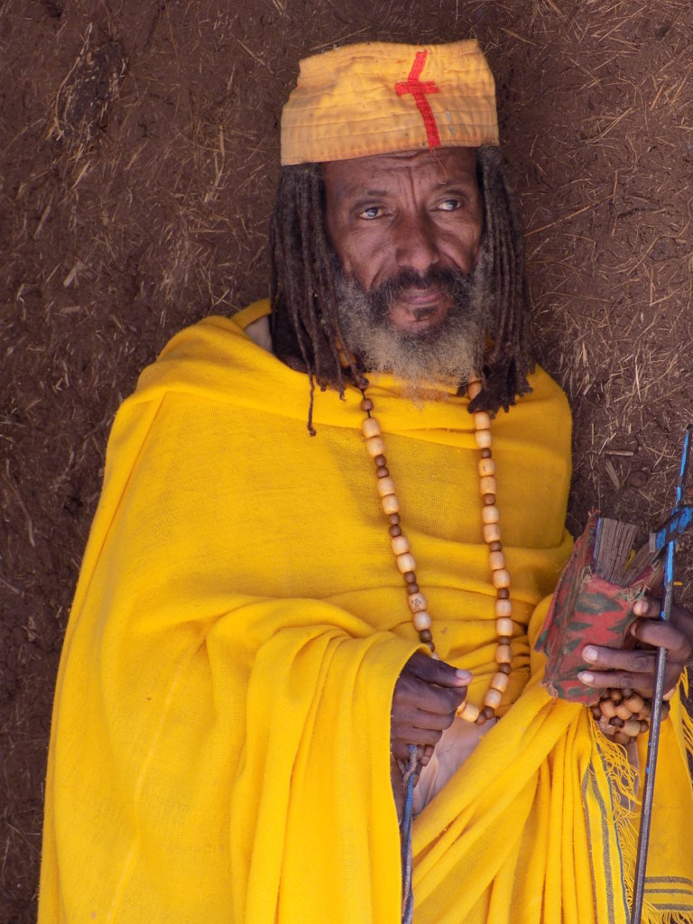This photo shows a priest in mustard yellow robes sitting outside Bete Maryam