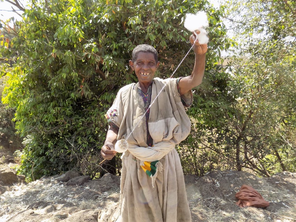 This photo shows a smiling toothless lady hand-spinning some wool