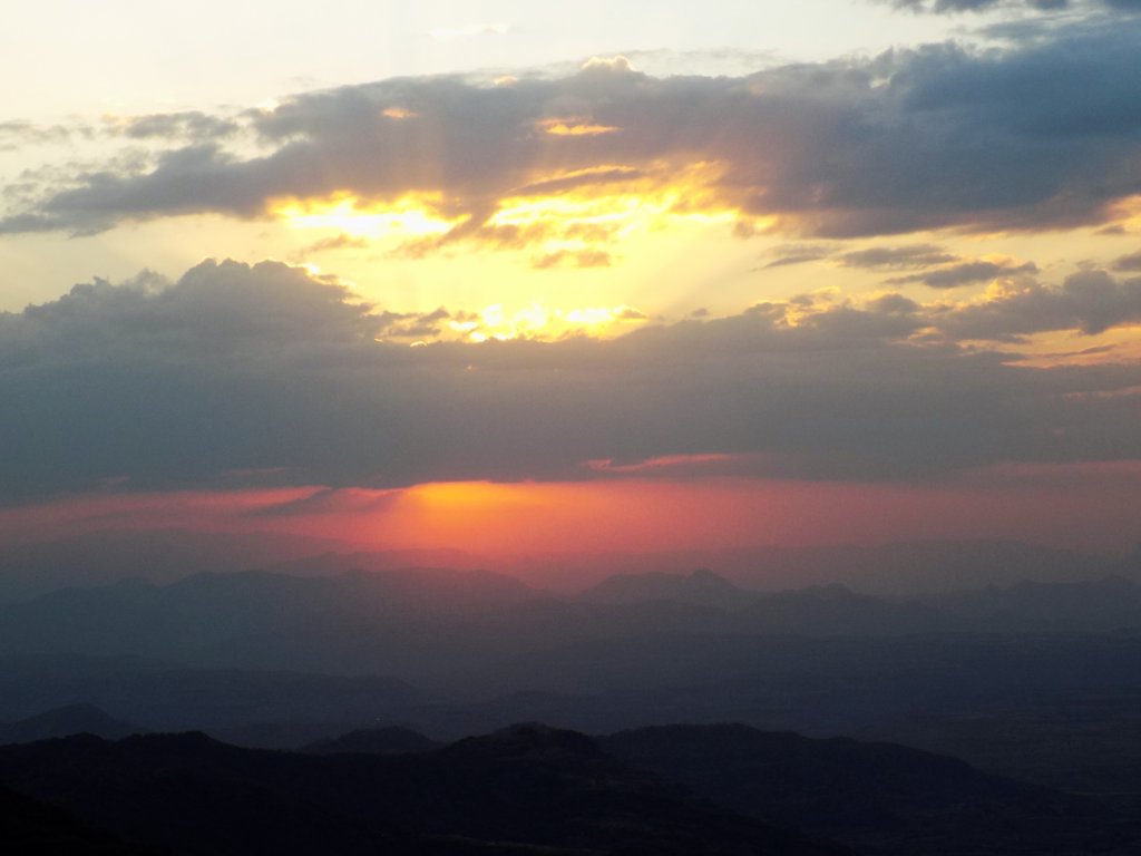 This photo shows a cloudy sky with the sun setting between the clouds and casting beautiful orange and yellow light