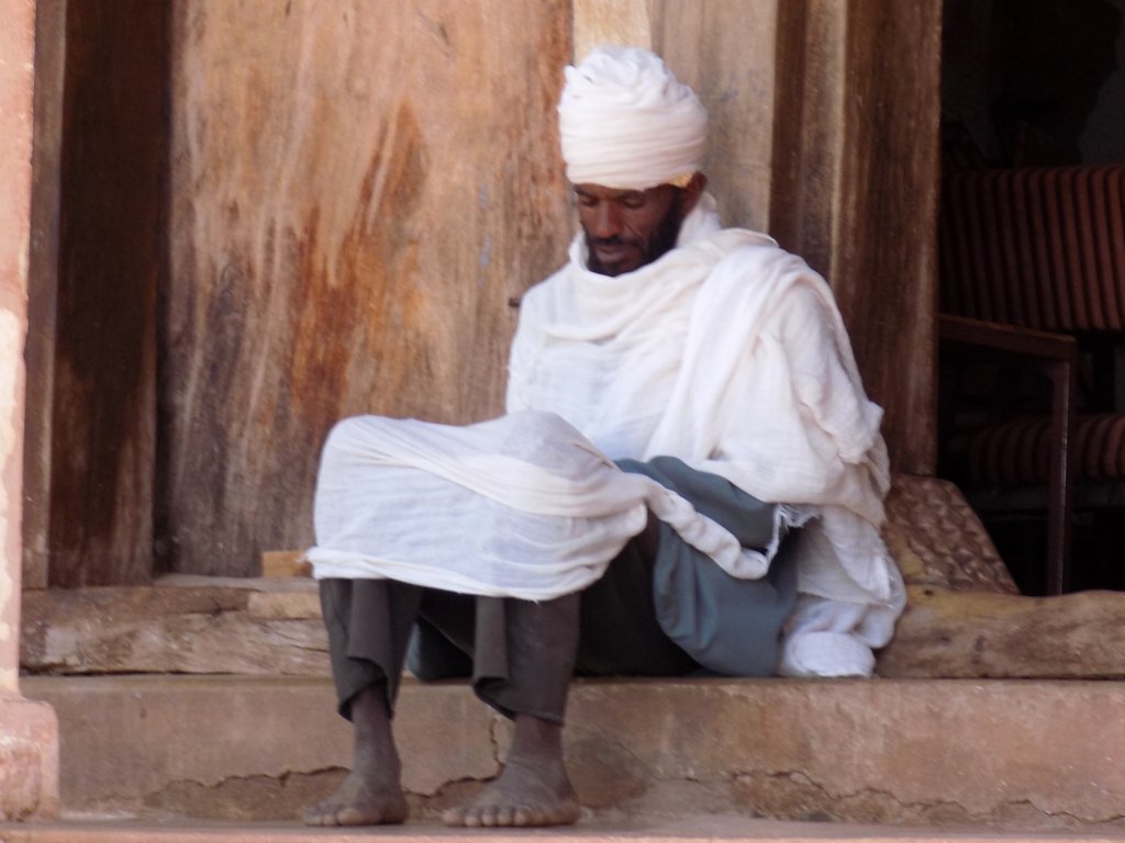 This photo shows a priest in his white robes sitting on the steps at the entrance to the church reading the Bible