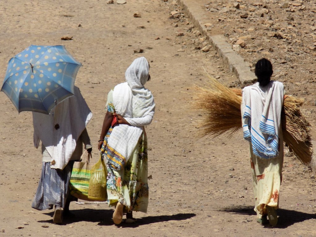 This photo shows the back views of three ladies in traditional dress walking away from the market laden down with their purchases