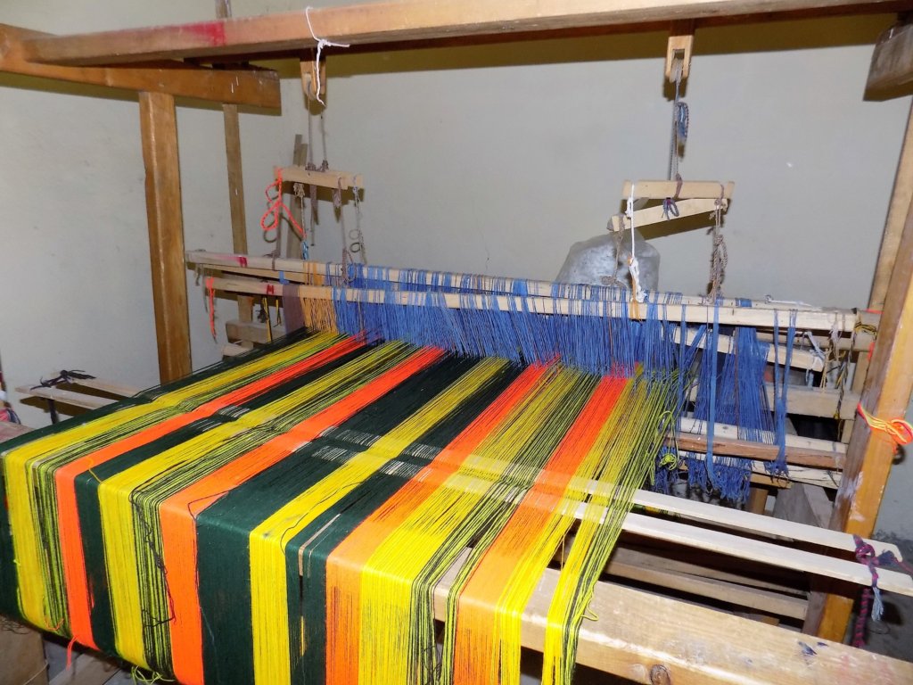 This photo shows orange, yellow and green wool being woven into fabric on a wooden hand loom.