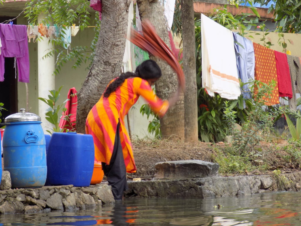 This photo shows a lady doing her washing in a backwater. She is bashing an item of clothing on a stone to get it clean