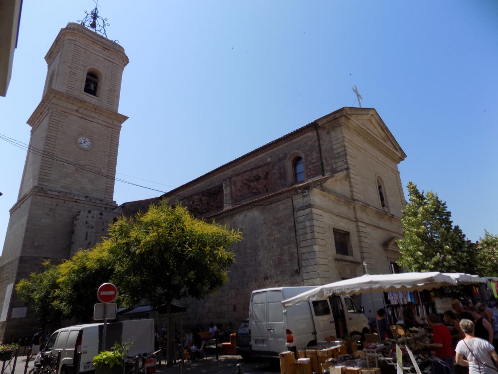 This photo shows Marseillan Church against the backdrop of a brilliant blue sky with a colourful market stall in the foreground.