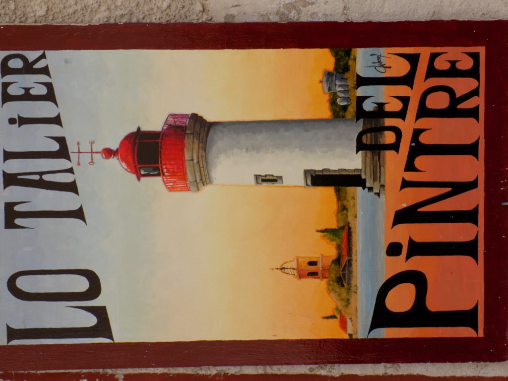 This photo shows a hand-painted sign displayed outside an artist's studio in Marseillan. It shows a lighthouse and harbour.