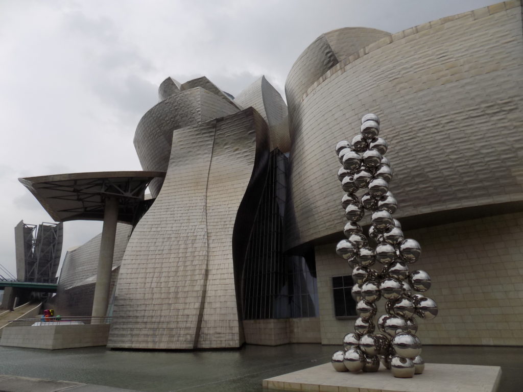 This photo shows the outside of the Guggenheim Museum with a modern art sculpture made up of large silver spheres piled on top of each other.