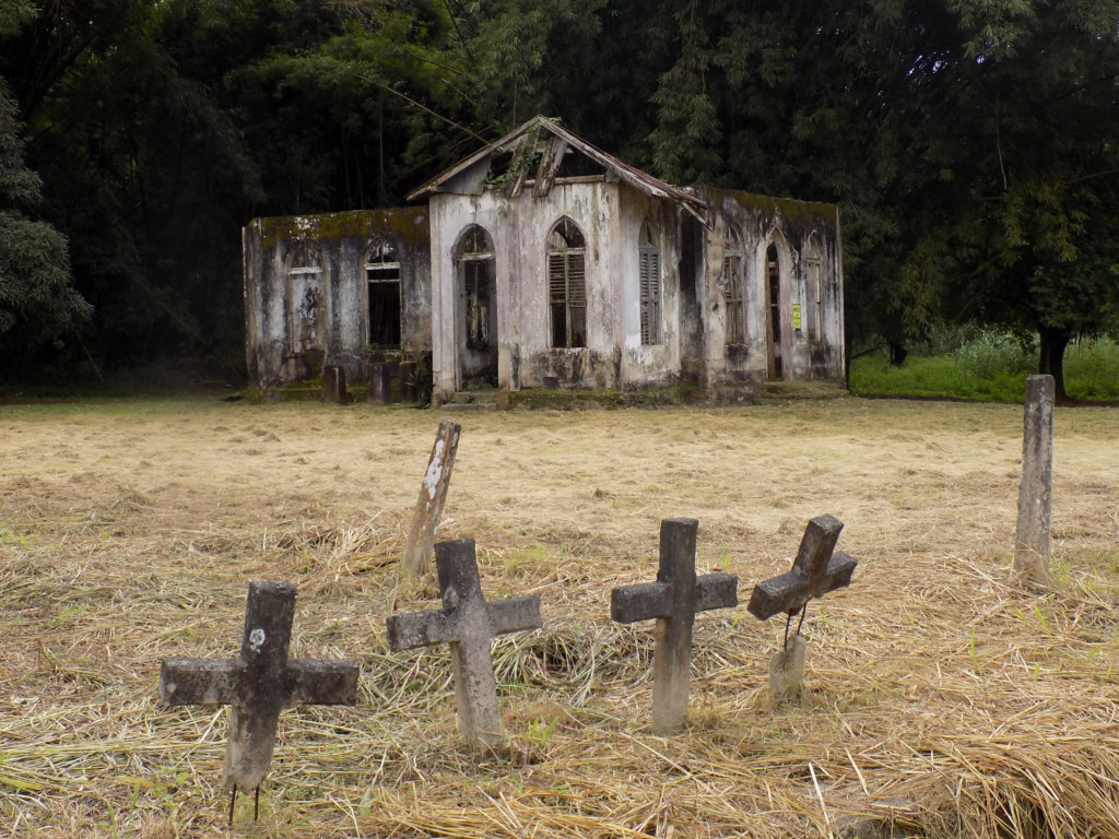 The image shows the ruins of St. Chad's Church, Mount St. Pleasant village, Trinidad. In the foregraund are a number of graves marked with simple stone crosses.