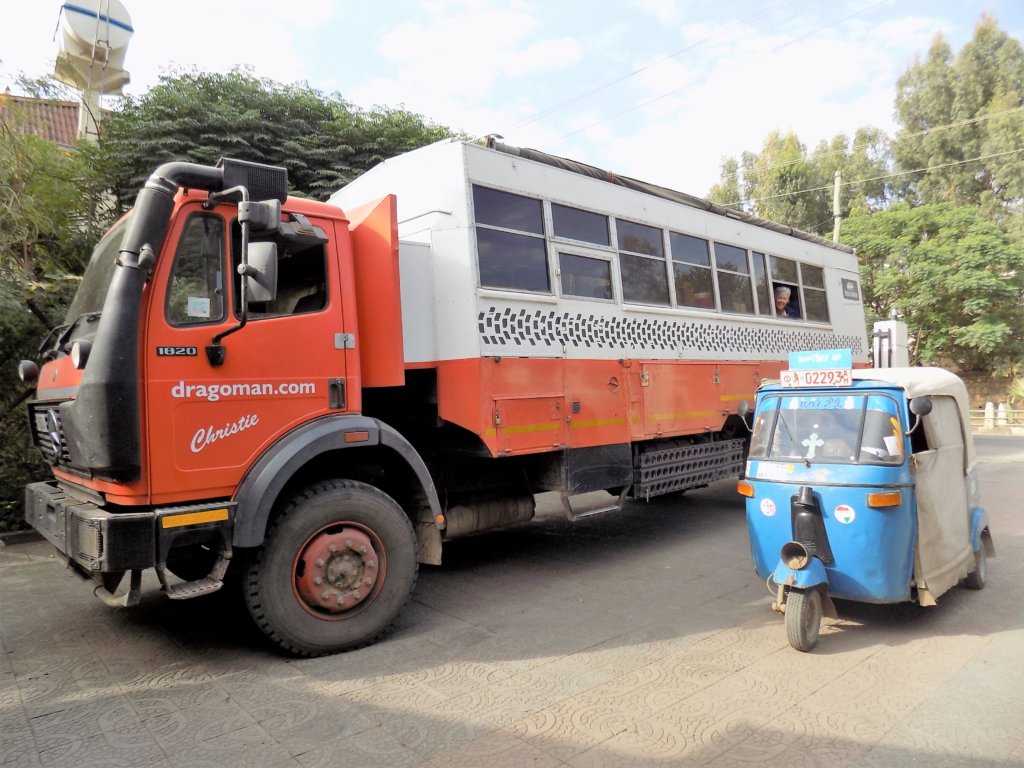 This picture shows a Dragoman truck parked next to a small, blue tuk-tuk
