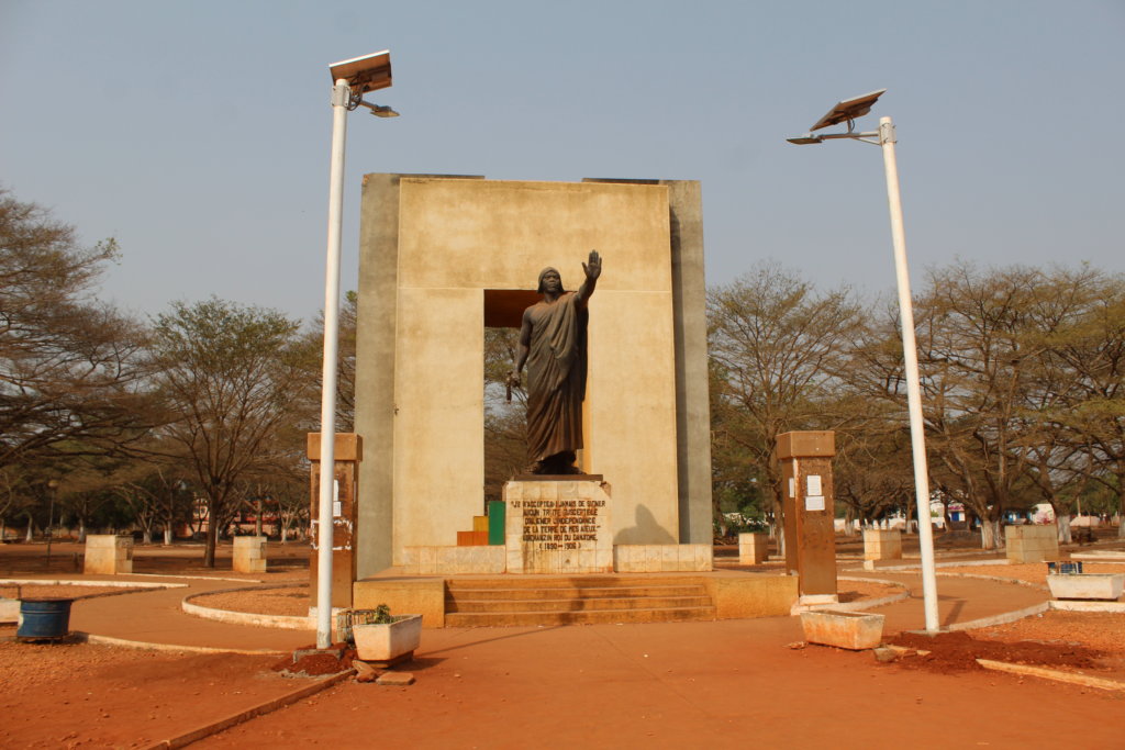 This photo shows a huge monument with a statue of King Gbehanzin in traditional dress in front of it