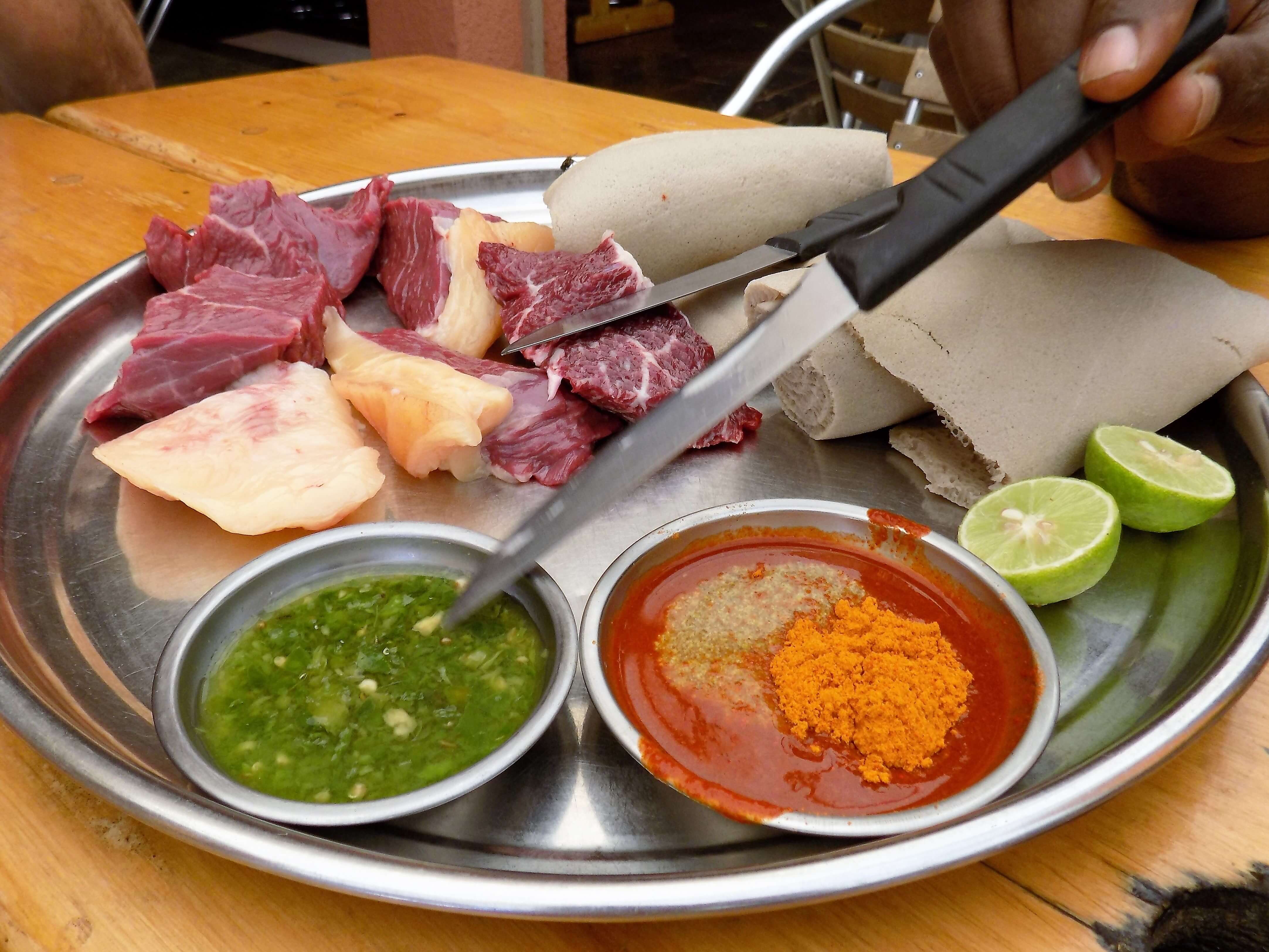 This photo shows a plate of raw beef, injera, limes and chilli sauces