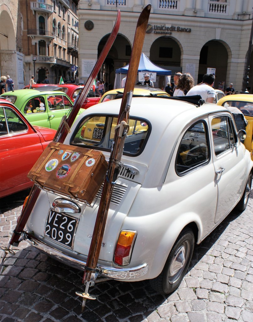 This photo shows a white Fiat 500 with an old-fashioned suitcase and a pair of wooden skis strapped to the back