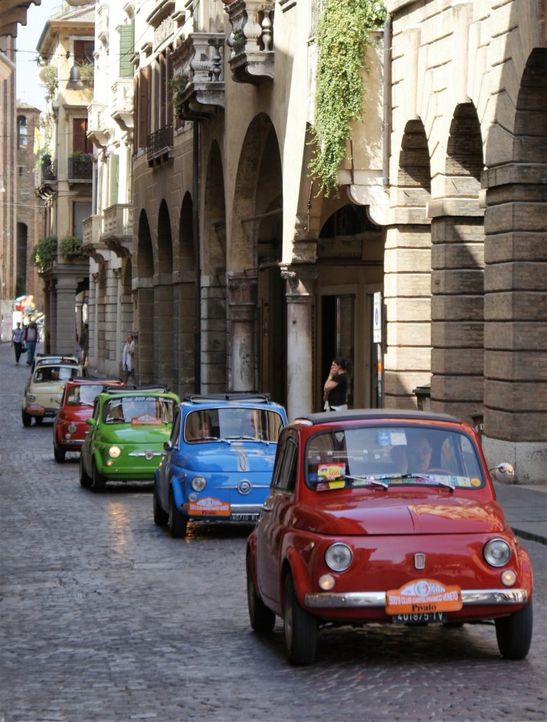 This photo shows a parade of brightly coloured Fiat 500s driving down an old street in Treviso