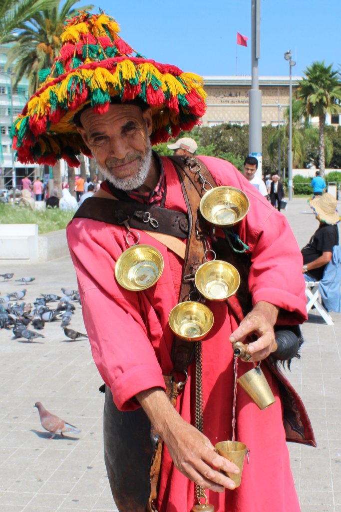 This photo shows a water seller in traditional dress with a bright pink coat and a multi-coloured wide-brimmed hat