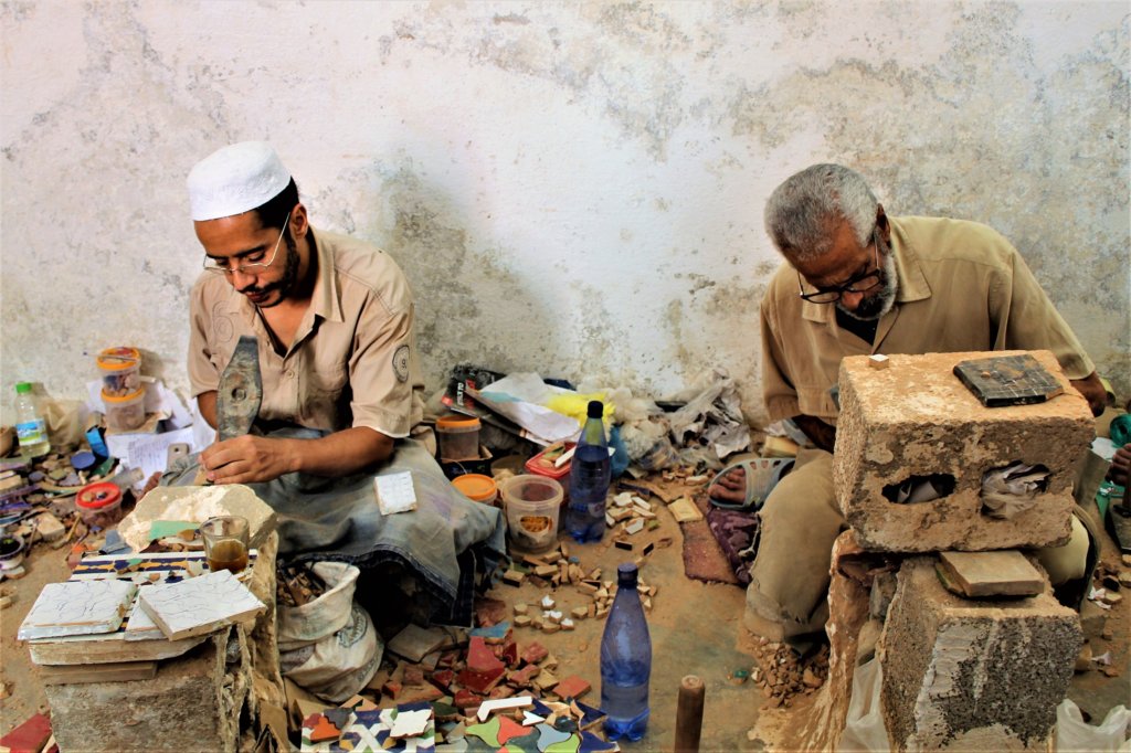 This photo shows two men sitting on low stools working on mosaic tiles