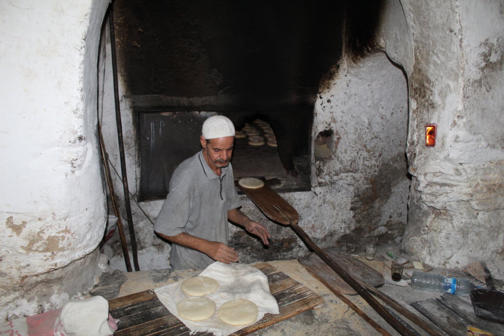 This photo shows a baker in a sunken oven loading bread with long wooden paddles 