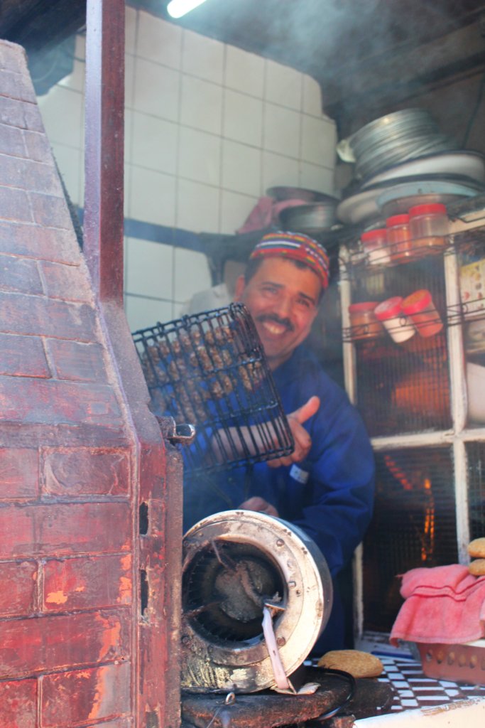 This photo shows a smiling man holding a grill full of camel burgers ready to go on the barbecue