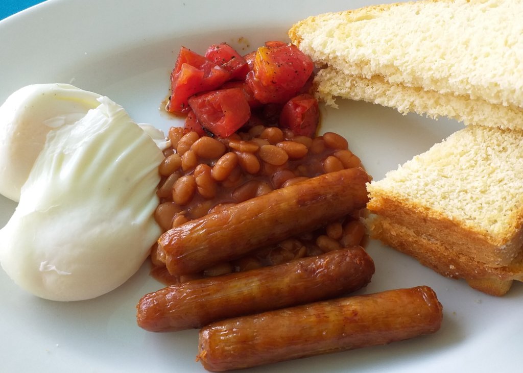 This photo shows a white plate with 3 thin pork sausages, some baked beans, two perfectly poached eggs, some roasted tomatoes, and 2 thick slices of white toast cut diagonally