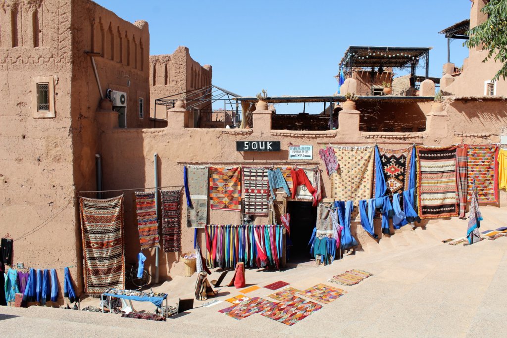This photo shows a terracotta building, the front of which is adorned with colourfu rugs and clothing for sale