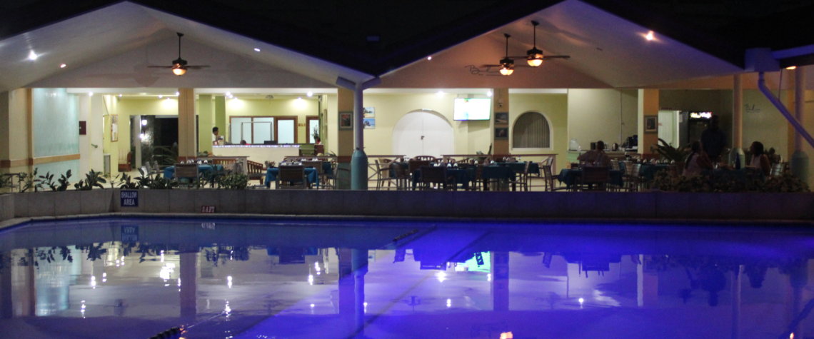 This photo shows the pool at night illuminated from underneath so that the water looks purple and you can see the restaurant reflected in it.