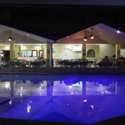 This photo shows the pool at night illuminated from underneath so that the water looks purple and you can see the restaurant reflected in it.