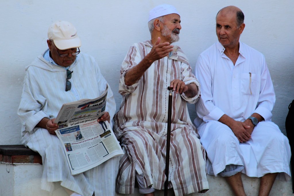 This photo shows three men sitting in the main square in Chefchaouen. One is reading a newspaper. The other two are sharing a story.