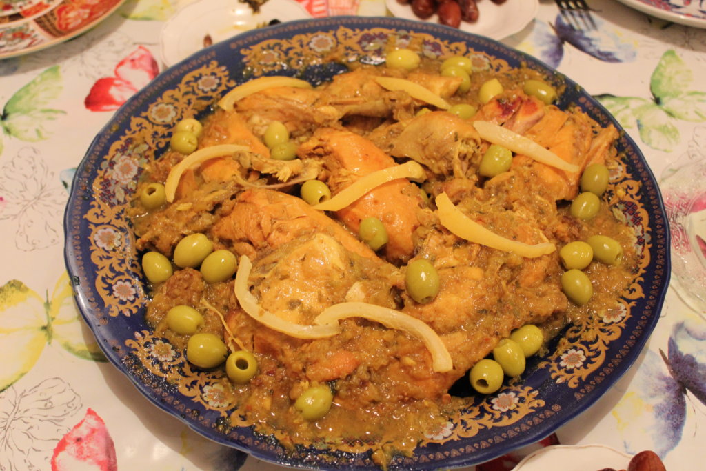 This photo shows a plate of chicken pieces in a rich yellow sauce garnished with preserved lemon slices and plump gree olives
