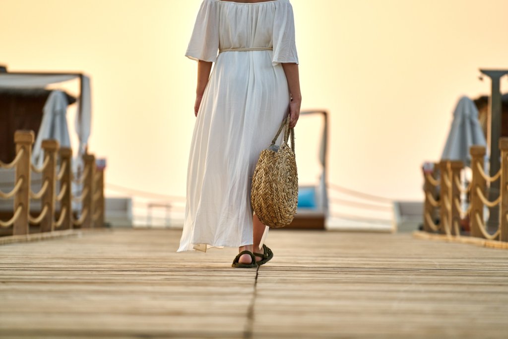 This photo shows a lady walking away from the camera wearing a long loose off-white dress and carrying a wicker bag
