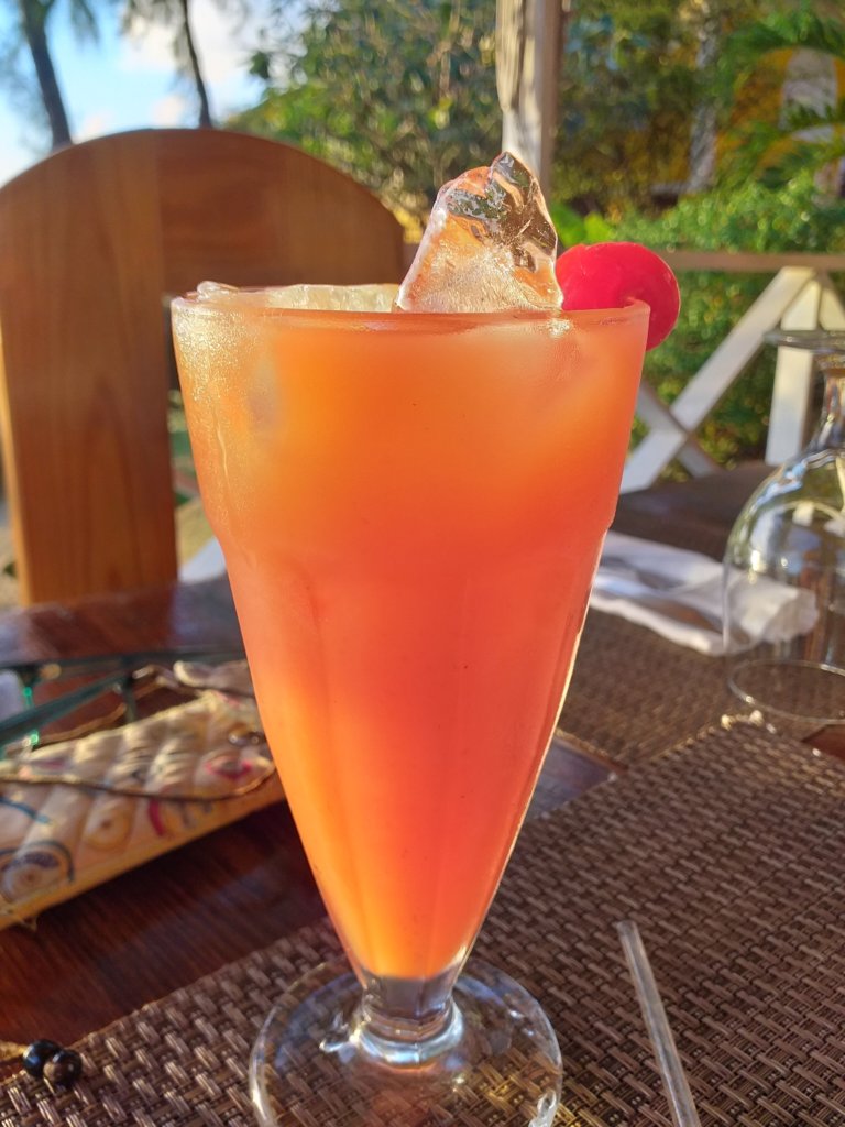 This photo shows a tall glass of orange liquid topped with cubes of ice and a cherry