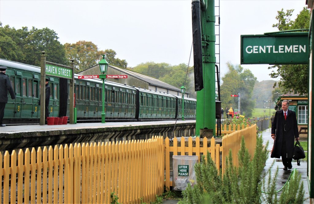 This photo shows several dark green railway carriages standing on a track behind a yellow picket fence