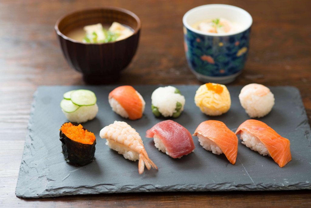 This photo shows a piece of black slate with 10 colourful pieces of sushi and 2 bowls of dipping sauces