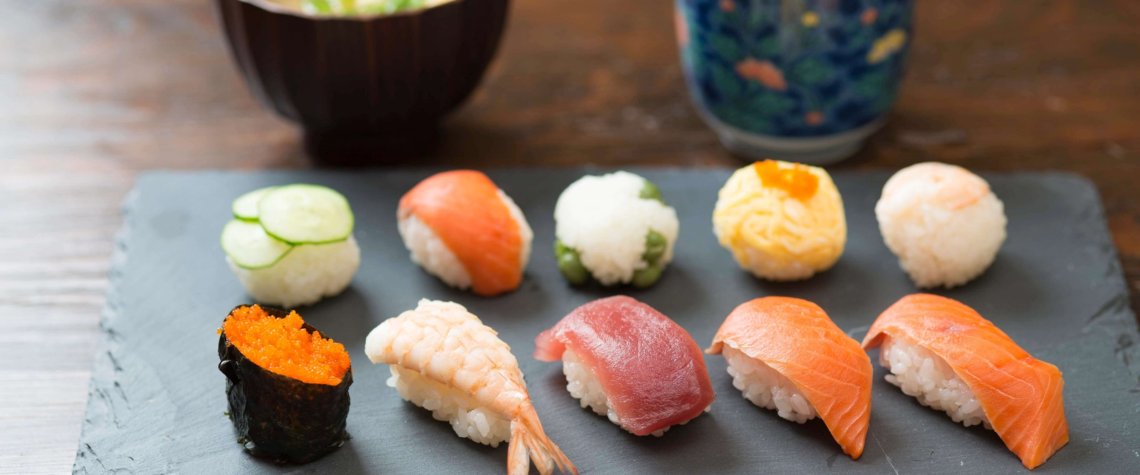 This photo shows a piece of black slate with 10 colourful pieces of sushi and 2 bowls of dipping sauces