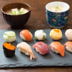 This photo shows a piece of black slate with 10 colourful pieces of sushi and 2 bowls of dipping sauces