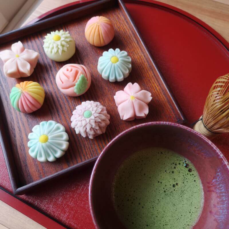 This photo shows a square plate with 9 pretty desserts in pastel colours