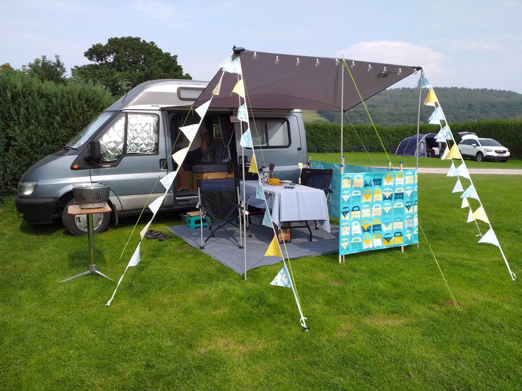 This photo shows our blue-grey campervan with its sun canopy out decorated with blue and yellow bunting