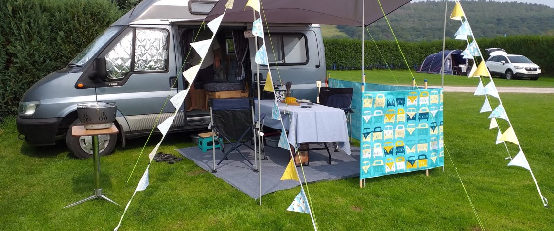 This photo shows our blue-grey campervan with its sun canopy out decorated with blue and yellow bunting