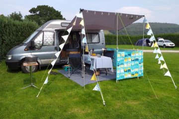 This photo shows our blue-grey campervan with its sun canopy out decorated with blue and yellow bunting