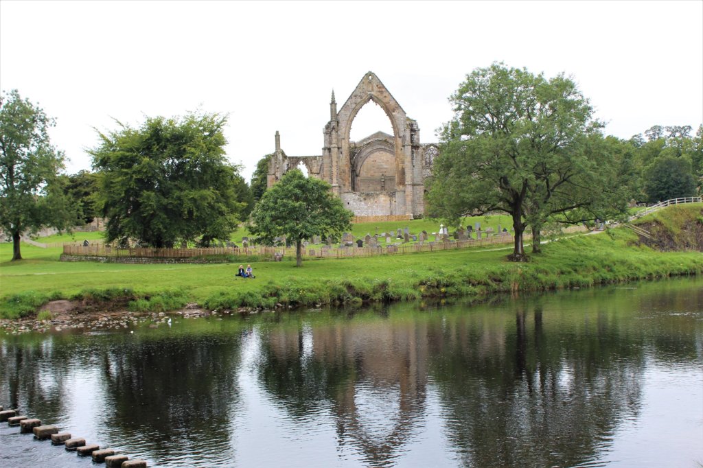 This photo shows the ruins of Bolton Priory reflected in the river with a series of stepping stones used to cross the wide river in the foreground