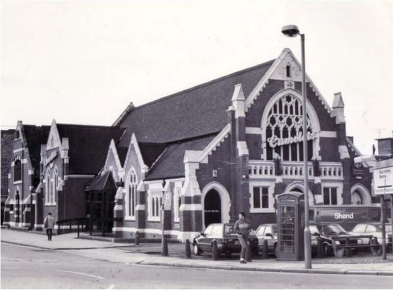 This black and white photo shows a traditional looking church with a Camelot's neon sign attached to the side