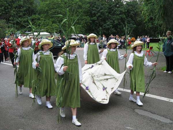 This photo shows several girls in green smock dresses and straw boaters carrying a sheet decorated with rushes and flowers