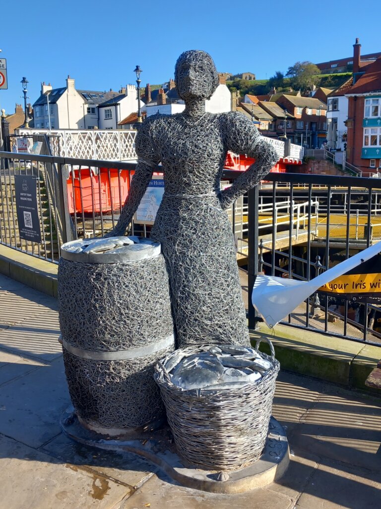 This image shows a wire sculpture of a fishwife standing against some railings with a barrel of fish
