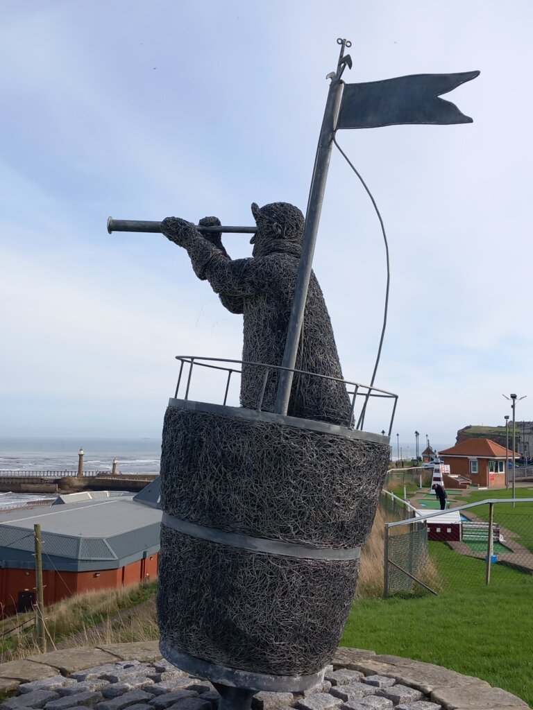 This image shows a sculpture of a man standing in a crows nest looking out to sea through a telescope