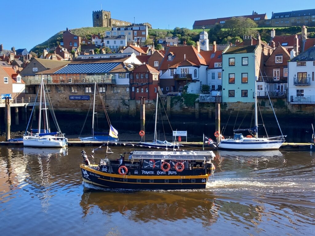 This photo shows a tourist boat leaving Whitby harbour with East Cliff and St Mary's Church in the background