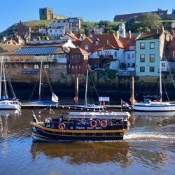 This photo shows a tourist boat leaving Whitby harbour with East Cliff and St Mary's Church in the background