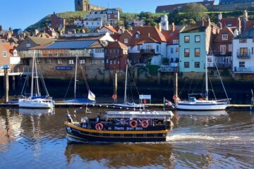 This photo shows a tourist boat leaving Whitby harbour with East Cliff and St Mary's Church in the background