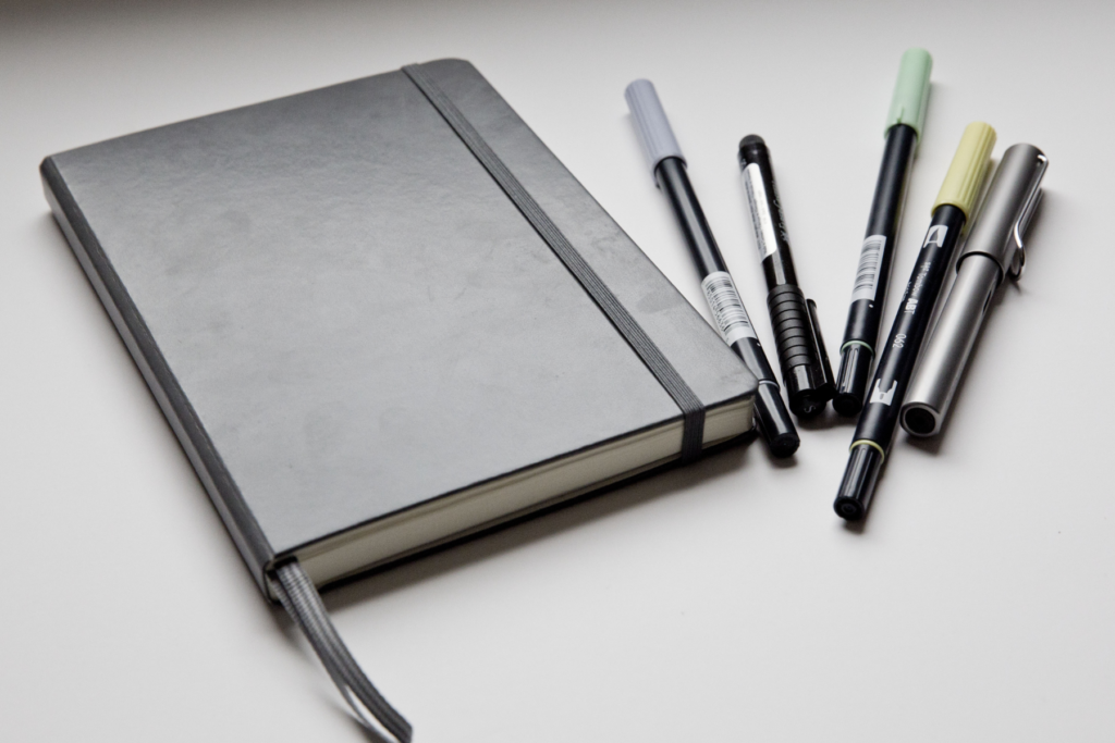This photo shows an A5 black notebook with ribbon markers and several pens at the side of it.